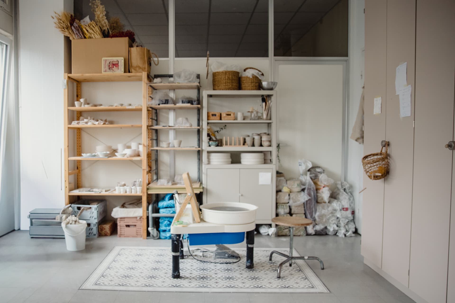 Atelier im Studio Sheree-Lou in Zürich-Altstetten: Töpferscheibe und Holzstuhl auf einer gemusterten Matte, dahinter Regale mit Keramik und verpacktem Ton.